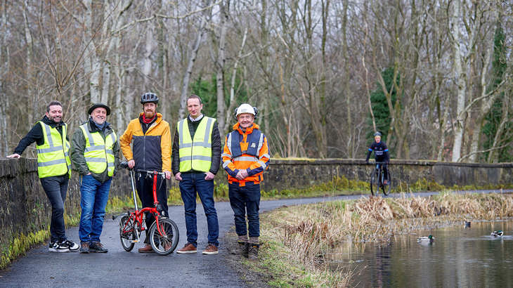 A small group of Walk Wheel Cycle Trust and Scottish Canals staff pose with members of the local community on the Monkland Canal towpath. A cyclist approaches in the background.