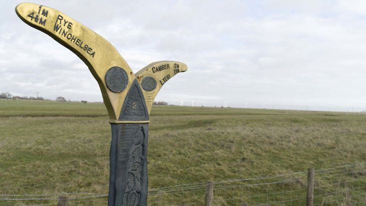 milepost marker on National Cycle Network route 2 showing the directions and distances from Rye and Camber, fields and windfarm in background. 