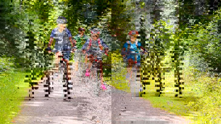 Three children on bikes on a traffic-free path of the National Cycle Network. Their grown up cycles behind. The path is surrounded by trees in full leaf. It is a warm, bright, sunny day.