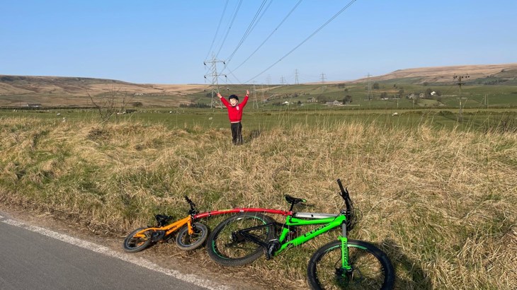 Little boy with his hands up in the air above his head. Stood in a field with his tandem bike in the foreground.