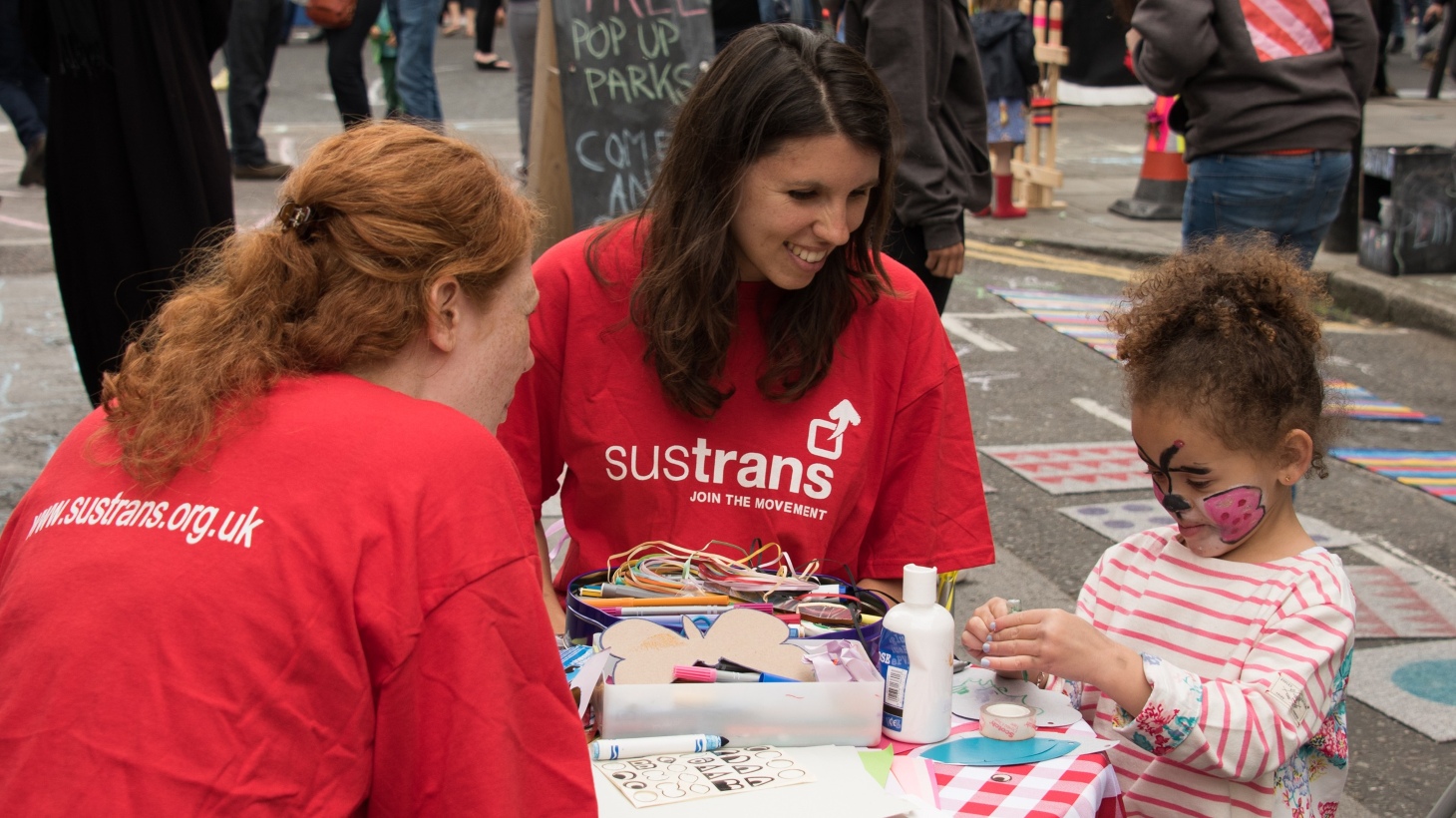Two volunteers sitting at a craft table at an event helping a little girl to cut and stick things 