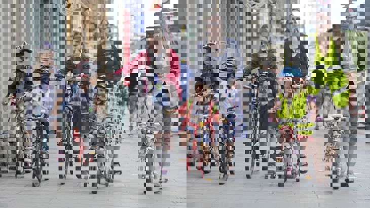 Young children cycling with adults walking on wide pavement