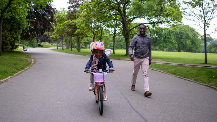 Young child cycling as her dad walks alongside her through a park
