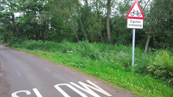A triangular warning road sign which says 'Cycles crossing' is beside a rural looking lane with SLOW painted in white on the surface of the road. The background is grassy with shrubs and trees behind.