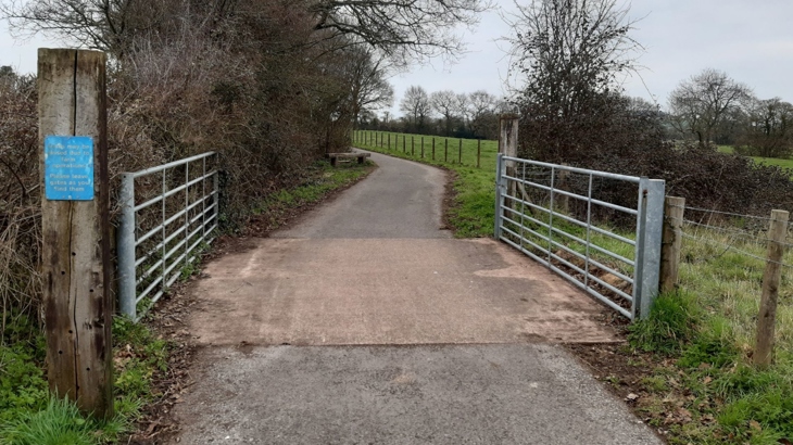 View of a traffic free path with two galvanized metal gates either side in the open position. An established hedgerow with trees appears to run to the left of the path beyond the gates and to the right, a fence with farmland beyond. The trees are bare, it appears to be winter.