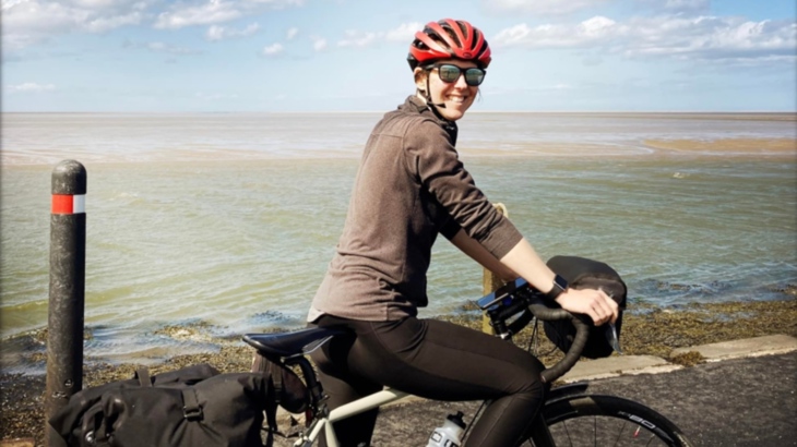 A woman riding a bike with panniers on a promenade on a sunny day with the sea in the background