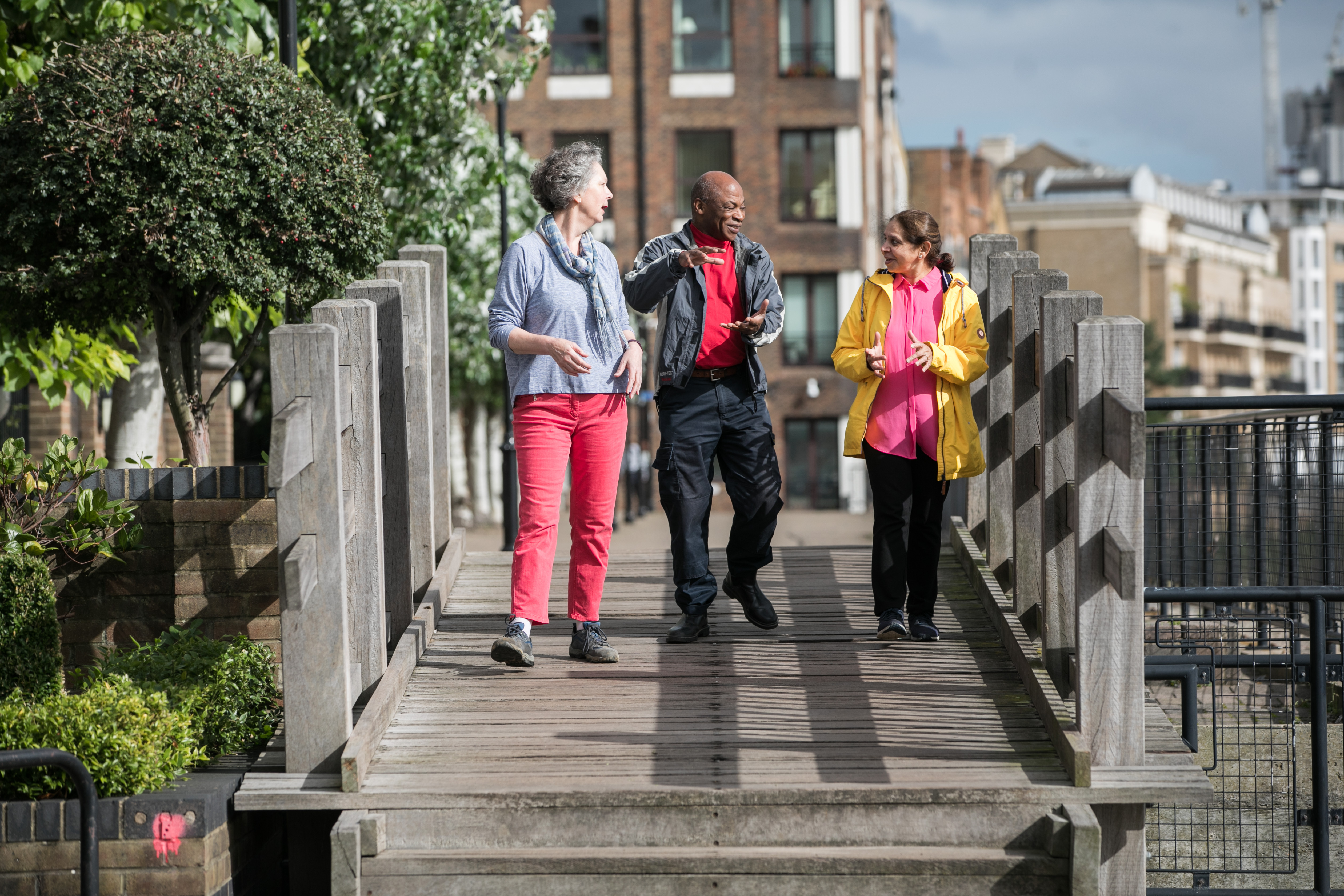 Three people are in conversation as they walk over a small wooden bridge in Tower Hamlets, London. A leafy green tree is to the left of them and tall buildings are in the distance.