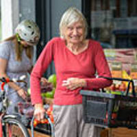 An older woman stood smiling wearing a coral long sleeved top outside of a fruit and vegetable market with a shopping basket hooked under her arm