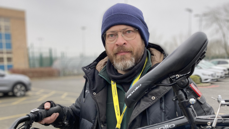 A man with a beard wearing thin framed glasses, a blue wooly hat and a warm looking coat holding a bicycle 