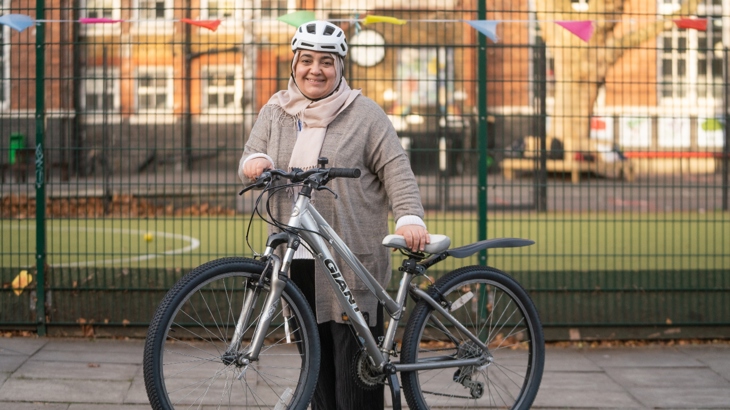 A woman wearing a headscarf under a cycle helmet stands in a school playground holding a bicycle. She is smiling, the autumn sun is shining, the mood is happy. 