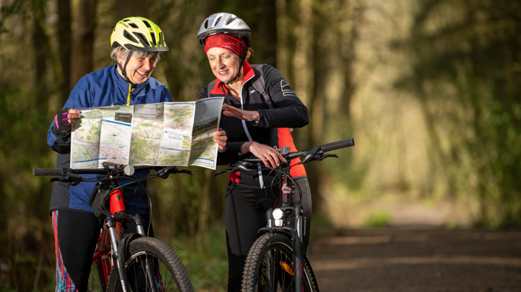 Two women are stopped on their mountain bikes on a traffic-free path in a woodland. They are looking at a map and smiling. They are wearing helmets and cycling clothing.