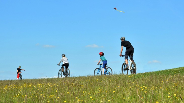A family cycling over the ridge of a hill in the Chilterns, with another playing with a kite against a clear blue sky