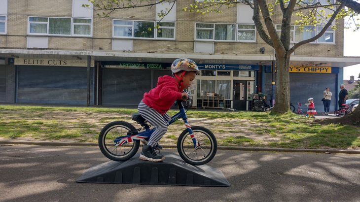 A young boy riding a bike over a small ramp in front of a row of shops in a residential area