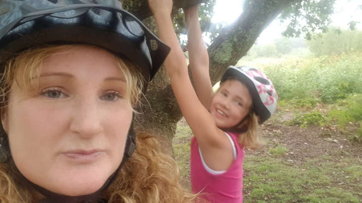 A woman wearing a bike helmet stood next to her daughter who is swinging on a tree branch also wearing a bike helmet