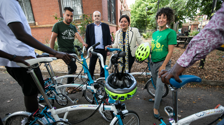 Four people stand in front of five blue and white Brompton bikes. In the foreground, other people stand holding bikes, their faces are obscured. The group are stood on a path, behind them are picnic benches and a red brick building. The day is bright and dry.