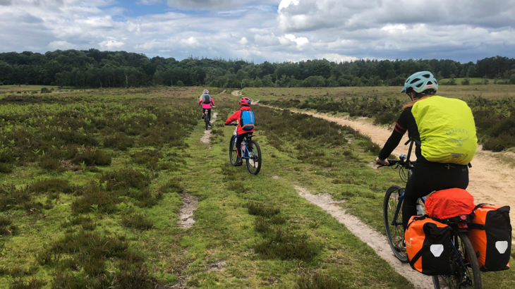 View from behind of a woman and two children cycling on a bridleway across a wild, rough green space towards a woodland. The woman is wearing a large yellow backpack and her bike has two orange panniers and a further bag. Ahead of her, the children each have a small backpack and helmet. The weather looks mild and the sky is blue with clouds.