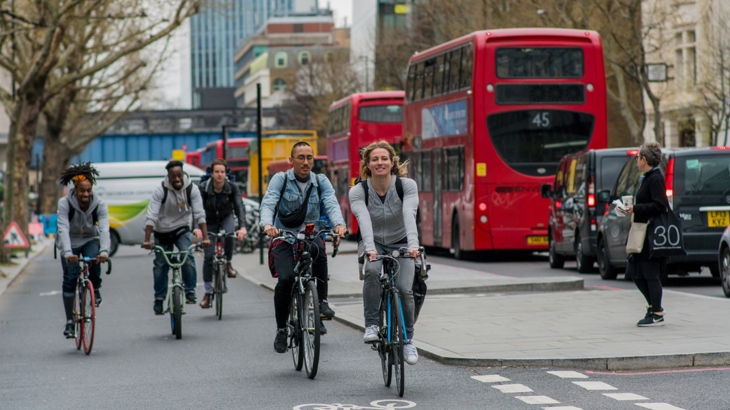 Five people cycling and smiling on Superhighway 6 in London. Three London red buses are in the background, along with tall buildings and a woman walking.