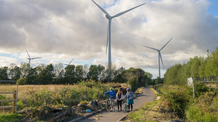 walking and pushing bicycles on a walking and cycling route that is due to be improved. wind turbines in the back ground, litter lines the route and the path surface is poor