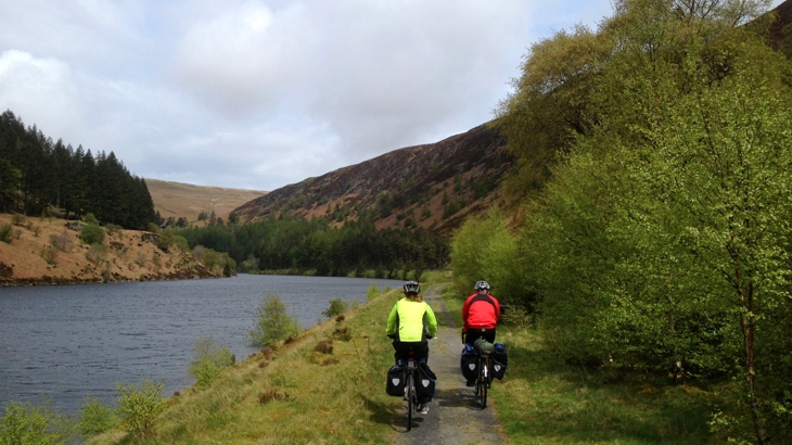 Two people riding bikes loaded with double pannier bags, cycle past a body of water in the Elan Valley, Wales. Beyond them are dramatic hills and woodland.