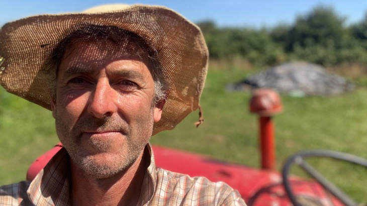Upclose shot of man's face, he is smiling and wearing a straw and stood in front of a red tractor.