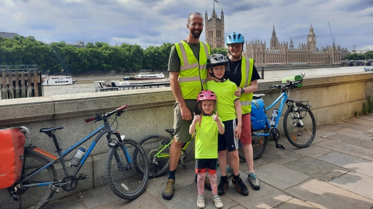 Emmer and her family pose for a photo in their helmets and cycling kit beside the river Thames in London. The House of Parliament can be seen across the water, beside them are their luggage-loaded bikes, and the group are all smiling.