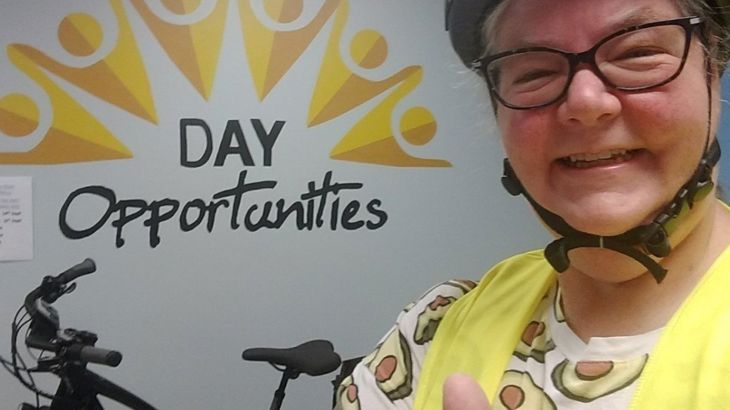 Sarah with glasses is wearing a helmet and high visibility vest and giving a thumbs up sign while standing beside an e-bike indoors at her workplace.