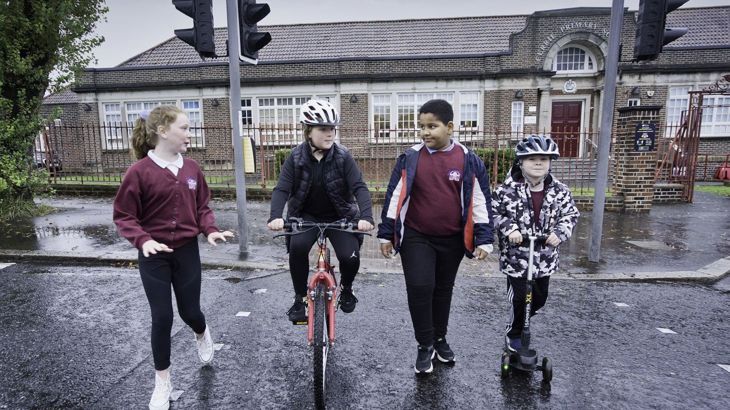 Four children are walking, scooting and wheeling at a crossing in front of Currie Primary School in Belfast.