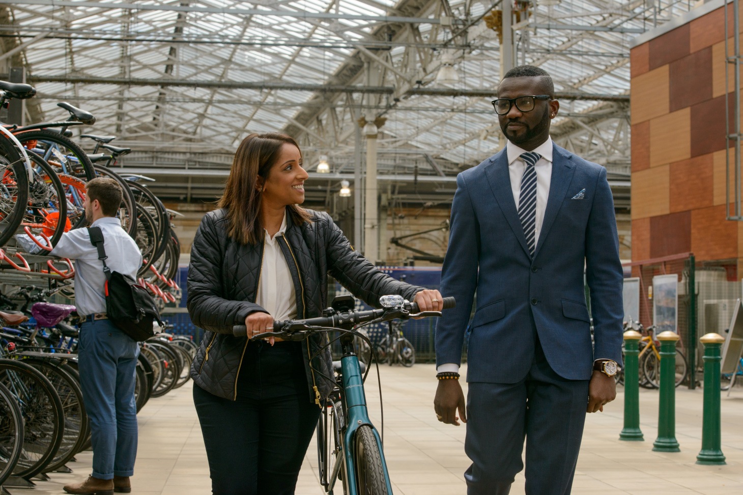 Two young professionals walking through a train station talking, one with a bicycle