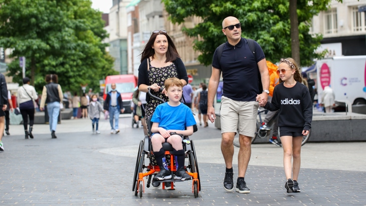 A man and a woman are walking in a pedestrianised shopping area. The man is holding the hand of a young girl and the woman is pushing a young boy in a wheelchair. The day looks dry and mild.