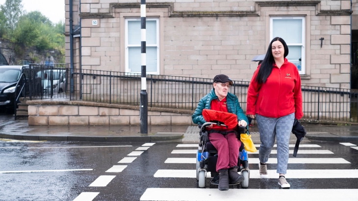 A man in a coat on a mobility scooter and a woman in a fleece, walking, are using a zebra crossing together. The road is wet and the day looks cool.