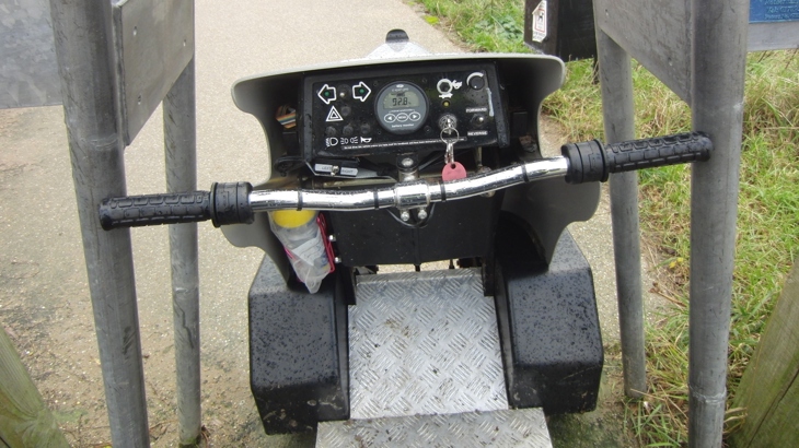 A rider point of view of a mobility scooter unable to pass through a metal 'A' shaped barrier on a traffic-free path.