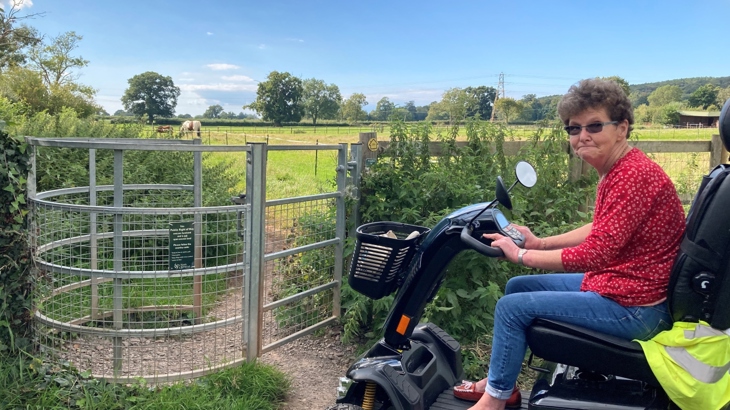 A woman riding a mobility scooter looks disappointed beside a metal kissing gate in the countryside which has a Public Right of Way notice attached to it. Beyond the gate is a beautiful green field, horses, trees and bright blue sky.
