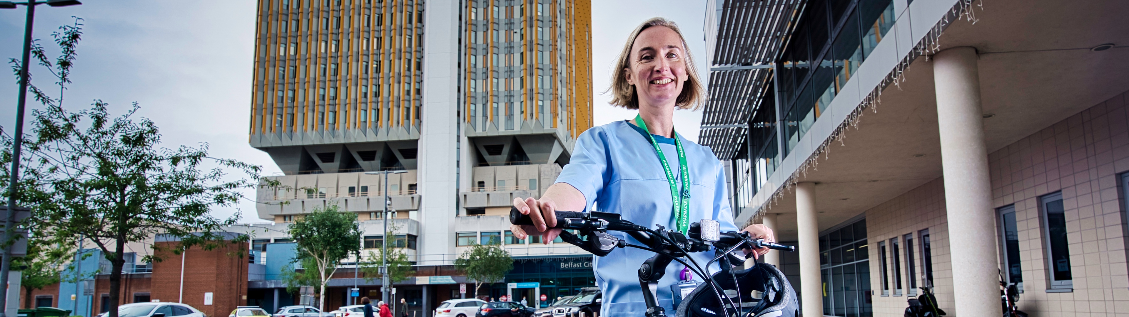 A female pharmacist in uniform and with a lanyard, stands outside a hospital in Belfast, holding the handlebars of her bike. 