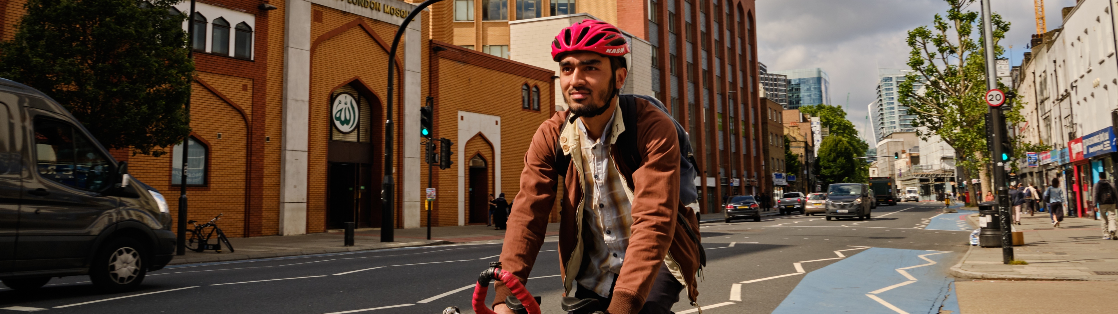 Man in a helmet, cycling in a bike lane on a busy street, past the East London Mosque. 