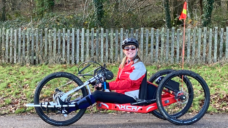 Laura sat smiling on her red XCR handcycle, on a traffic-free path. Behind Laura is a picket fence and green verge.