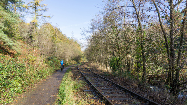 An image of NCN Route 884 - the image shows the route, which is surrounded by vegetation and some trees, on a sunny day with blue skies with a cyclist in the distance. There's also a disused railway path running alongside the cycle path.
