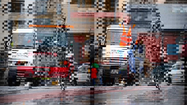 A man cycles in the rain in a city while wearing a high vis vest
