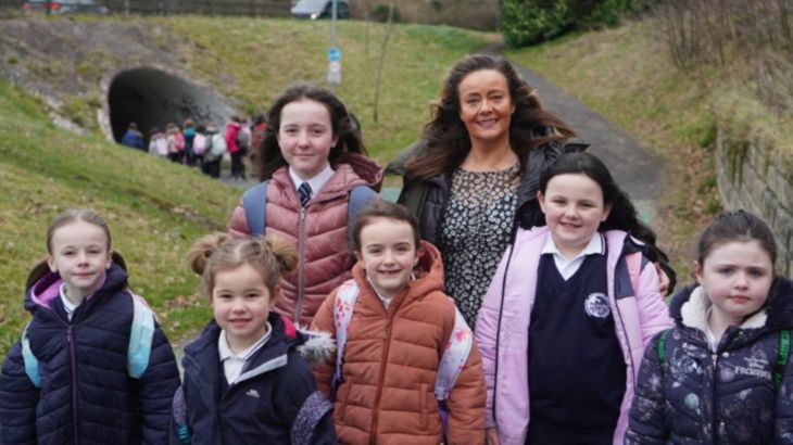 Six primary school girls stand outdoors with their female teacher smiling while a larger group of pupils walk through a pedestrian tunnel behind.