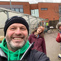 Walk Wheel Cycle Trust Walking and Cycling Technical Lead, Wayne Brewin, smiling and taking a selfie-style photo with two colleagues in front of bicycle storage in the school playground.