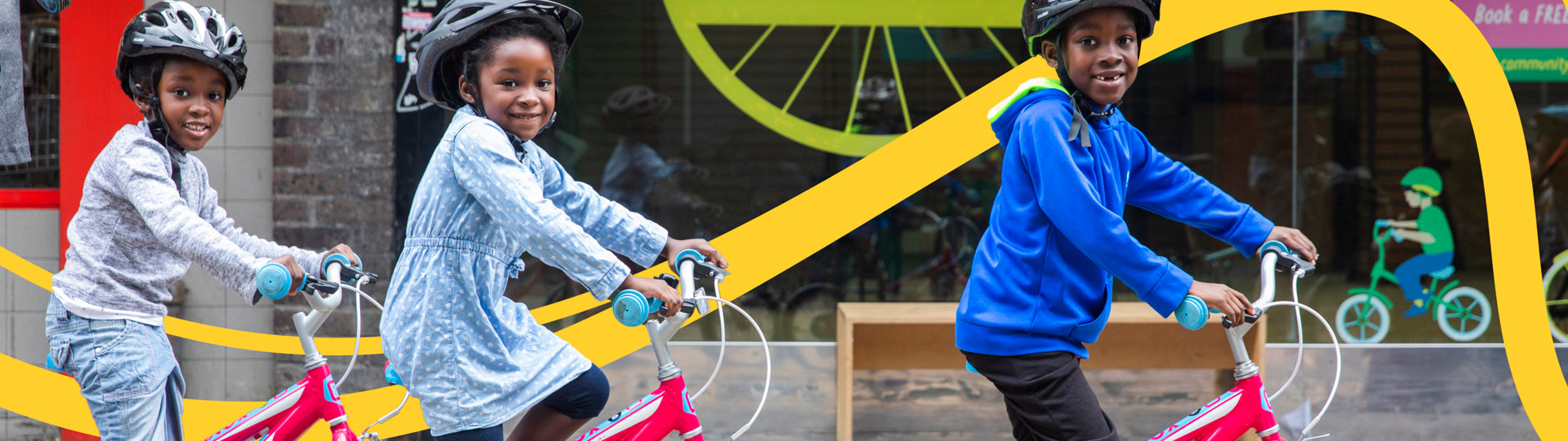 Three young children wearing helmets cycling outside of a bike hub in London, smiling and laughing as they follow each other.