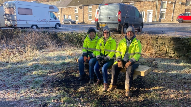 Three members of Ogmore Valley Priide, a local community group, sat on a bench in a residential area of Ogmore Valley. They're facing the camera, smiling, wearing fluorescent jackets in wintery sunlight.