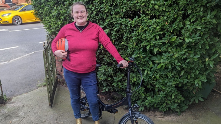 A young white woman smiling wearing a pink jumper stood with her electric foldable cycle outside of her home in Manchester