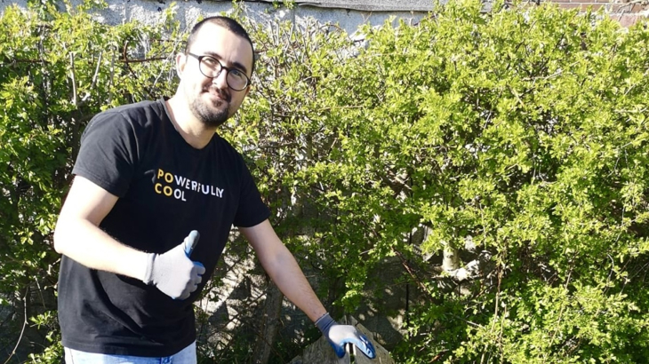 A man wearing glasses and a black t-shirt stood in front of a hedge giving a thumbs up while smiling