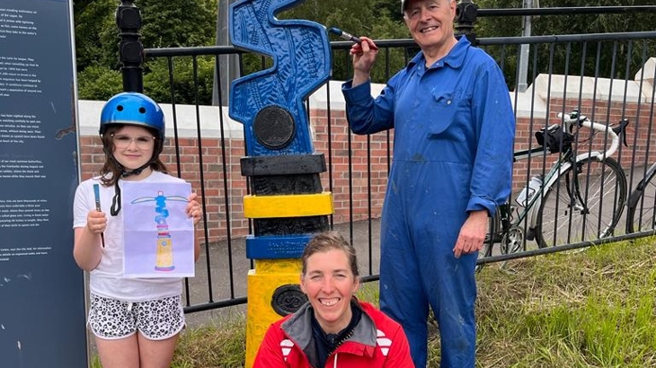 A man is standing beside a sculpture with a paintbrush while a woman crouches in front and a little girl stands to the other side holding a picture.