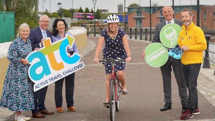A woman on a bicycle wearing a helmet smiles at the camera while a group of men and women stand on each side of her on a traffic-free, riverside path.