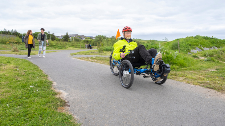 A man with a white beard wearing high-vis using an adapted trike in Arcadia Park in Scotland on a cloudy day