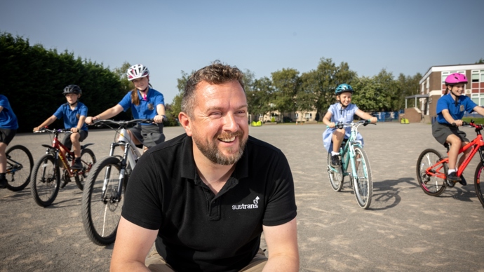 A man wearing a black Walk Wheel Cycle Trust top smiles at something off camera with children on bikes behind him in a school playground.