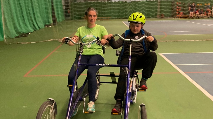 A parent and child riding an adaptive bicycle in an indoor setting for Ysgol Pendalar's sponsored walk and wheel.