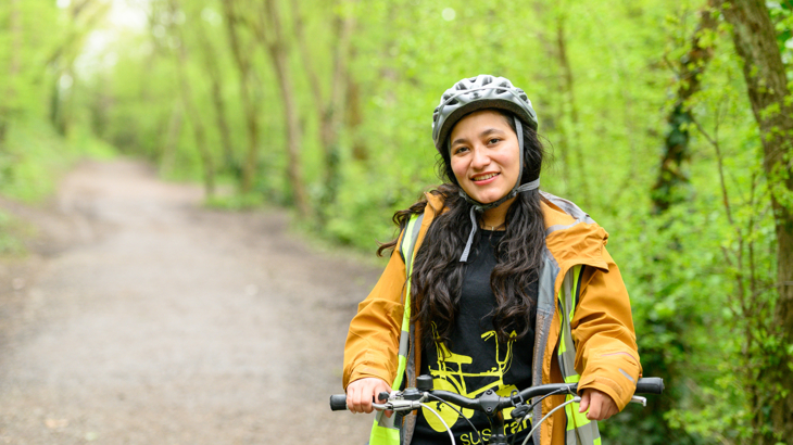 A young woman wearing a yellow rain coat and a helmet stood with her bike smiling in a woodland setting in England