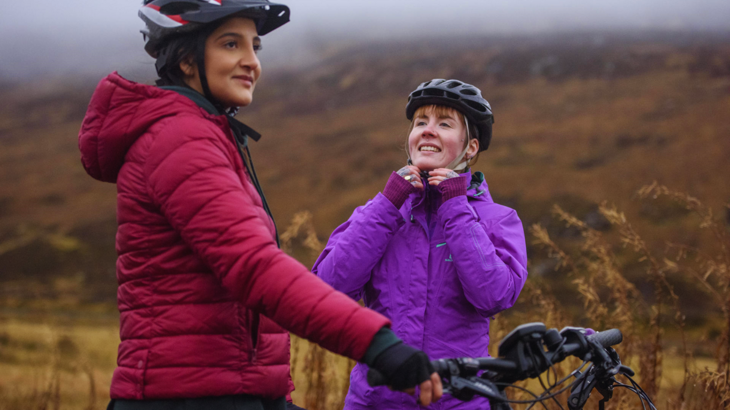 Two women wearing waterproof coats and cycling helmets on National Route 7 in the Scottish Highlands.